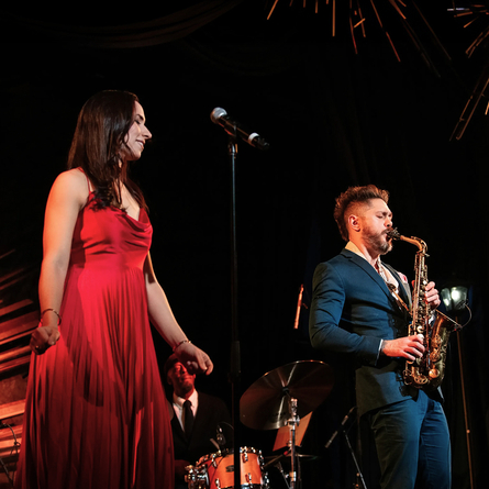 A woman in a red dress stands near a microphone, while a man in a blue suit plays a saxophone.