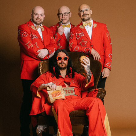 A photo of the band Elvana, they are clad in red, one of the singers in sitting on a chair holding a burger.