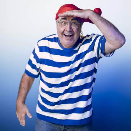 Danny Baker in a blue and white striped t-shirt and a red hat. He is holding his hand to his forehead