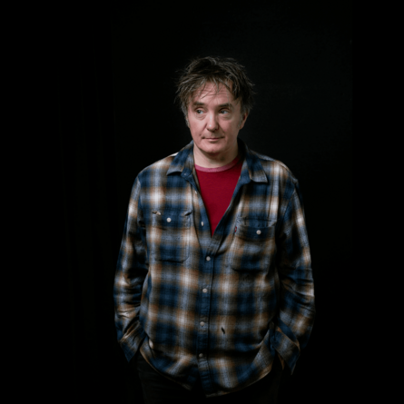 A photo of Dylan Moran, he is wearing a red shirt under a plaid button down shirt. He is standing against a dark background.